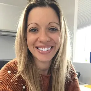 michelle has long blonde hair and is wearing an orange sweater sitting at a desk